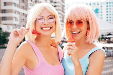 Two young beautiful smiling hipster female in trendy summer clothes. Sexy carefree women posing in the street in wigs. Positive funny models having fun at sunset. In sunglasses. Eating candy lollipop