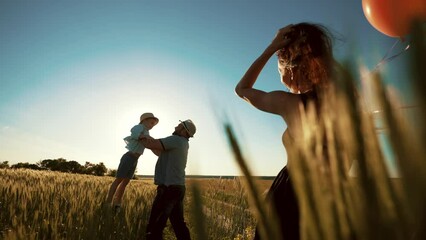 Mom is standing in field and her hair is beautifully fluttering in wind, holding balloons in hands and watching as dad lifts his son on arms and throws him up. Happy family weekend outing in country.