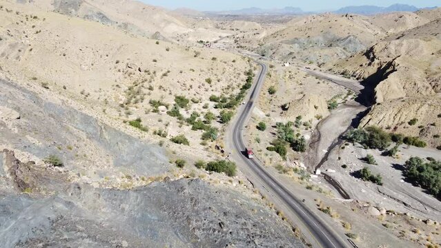 Aerial drone zoom in shot of vehicle driving along winding RCD Road surrounded by arid vegetation in Balochistan on a sunny day.