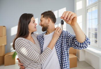 Happy young family couple embracing, showing keys to camera in living room. Excited loving spouses homeowners celebrating moving in new apartment. Moving day, real estate concept