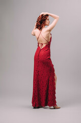 Young ginger woman dancing in a sexy lace red dress. Bright open-back maxi outfit, rear view. Full length backless evening gown with straps and split. Natural looking wavy hair. Valentine's day look