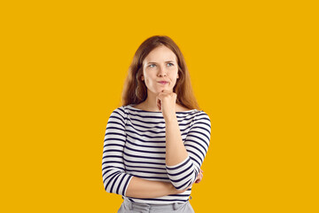 Portrait of serious confused undecided young woman full of doubt holding hand on chin and thinking, trying to make hard choice decision. Girl with wavy hair in striped sweatshirt on yellow background.