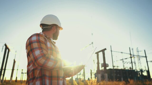 Design engineer in helmet holds laptop in hands at construction site in field, estimating investment attractiveness this object. Male compares his project with customer project. Using technologies.