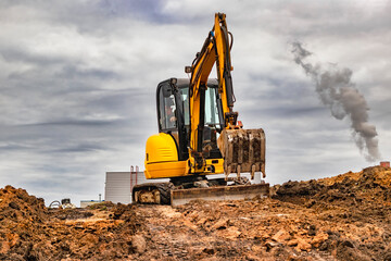 Mini excavator at the construction site on the edge of a pit against a cloudy blue sky. Compact...