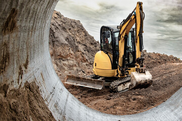 Mini excavator at the construction site on the edge of a pit against a cloudy blue sky. Compact...