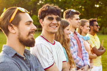 Cheerful young man standing with his friends in row holding hands during fun in summer park. Group of joyful multiracial young people having fun together outdoors. Selective focus. Cropped image.