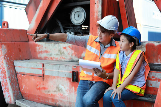 Asian foreman father and his daughter sitting on forklift while using computer and teaching little girl. Engineer dad pointing something with his kid at industrial container cargo freight ship.