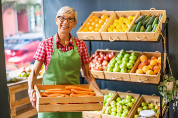 Mature woman works in fruits and vegetables shop. She is holding basket with carrot.