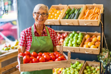 Mature woman works in fruits and vegetables shop. She is holding basket with tomatoes.