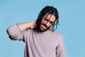 Fototapeta premium Arab man suffering from neck pain discomfort and massaging muscles with face grimace. Young adult person with black braided hair having painful cramp in back and spine problem