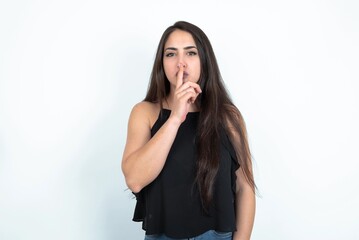 young brunette woman wearing black blouse over white studio background  makes silence gesture, keeps finger over lips. Silence and secret concept.