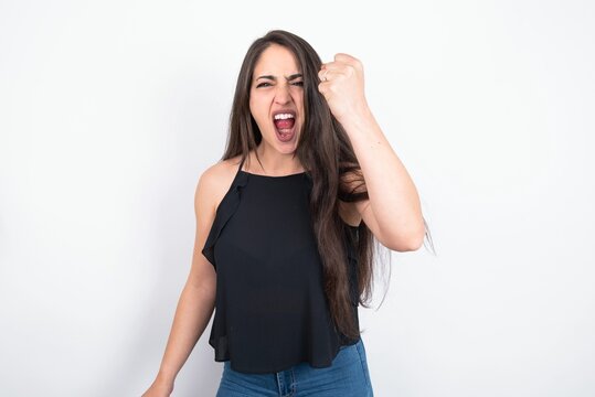 young brunette woman wearing black blouse over white studio background angry and mad raising fist frustrated and furious while shouting with anger. Rage and aggressive concept.