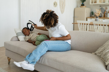 Caring black mother removes vr 3D glasses from tired little son fell asleep on couch in living room...