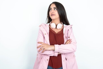 Pleased young brunette woman wearing pink raincoat over white studio background keeps hands crossed over chest looks happily aside