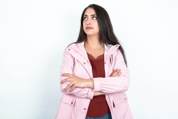 Charming thoughtful young brunette woman wearing pink raincoat over white studio background stands with arms folded concentrated somewhere with pensive expression thinks what to do