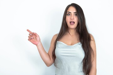 Emotive brunette woman wearing white tank top over white studio keeps jaw dropped from shock demonstrates amazing promo points right on blank space demonstrates big shopping sale. Advertisement