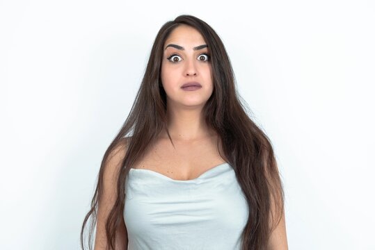 Stunned brunette woman wearing white tank top over white studio background stares reacts on shocking news. Astonished girl holds breath