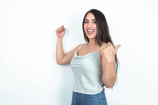 Hooray cool brunette woman wearing white tank top over white studio background point back empty space hand fist