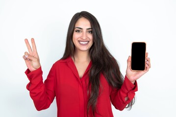 Fototapeta premium Young brunette woman wearing red shirt over white studio background holding modern device showing v-sign