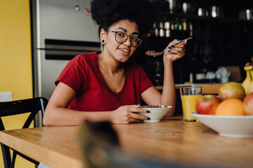Smiling african woman drinking orange juice and eating cereal while sitting in kitchen