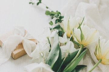 Obraz na płótnie Canvas Happy Mothers day. Beautiful white bouquet with gift box on rustic wooden table. Stylish spring tulips and daffodils on soft fabric and present, holiday still life. Womens day banner