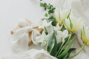 Beautiful white bouquet with gift box on rustic wooden table. Stylish spring tulips and daffodils on soft fabric and present, holiday still life. Happy Mothers day. Womens day banner