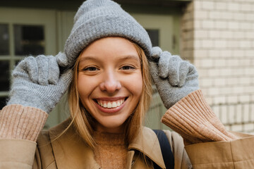 Portrait of woman adjusting warm hat and smiling at camera while standing outdoors