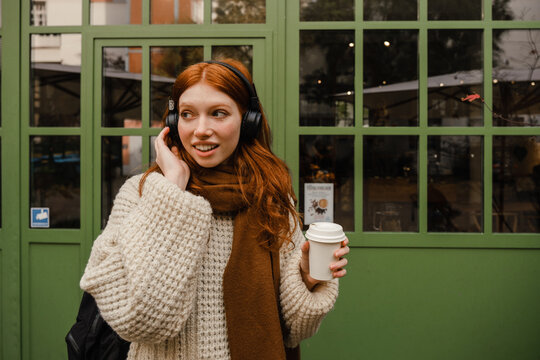 Smiling redhead woman listening music with headphones and drinking coffee while standing outdoors