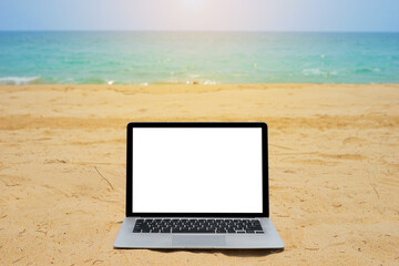Laptop with blank screen for creative design on sand beach nearby blue sea background with sun ray effect. Computer notebook with monitor clipping path for present landing page design mock up template