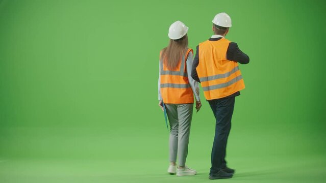 Green Screen.Female and Male Workers Partners in Safety Jackets and White Helmets,Holding Tablet and Paper Clipboard Walking Through Discussing Project Standing Back to Camera.Team Work Concept.