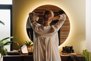 Back view of woman touching her hair while standing in front of mirror in bathroom
