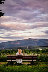 Naklejka na ściany i meble Young blonde woman sitting on wooden bench in park resting with beautiful mountain and sky view