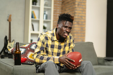 Young African man watching sport match on television and supporting his favorite team. Happy man enjoying victory, watching sports competition on tv.