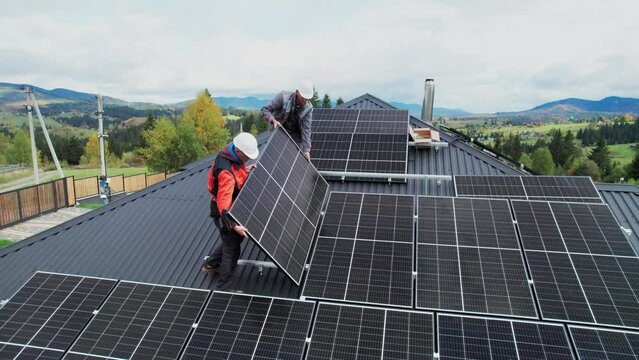 Technicians building solar panel system on roof of house. Drone view of men workers in helmets carrying photovoltaic solar module outdoors. Concept of alternative and renewable energy.