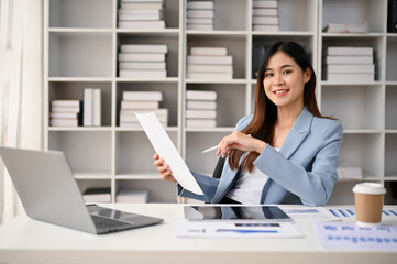 Confident Asian businesswoman sits at her desk in her private office with business reports.