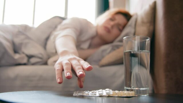 Glass of water and cure drugs on table in bedroom blurry sick woman suffering from headache in bed morning lady painful head migraine fever hangover pain flue coronavirus girl take pills painkiller
