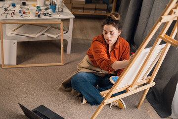 Female painter sitting with laptop in home art studio, having online art class. Top view.