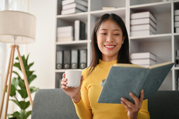 Young Asian woman reading book in her living room while drinking a coffee