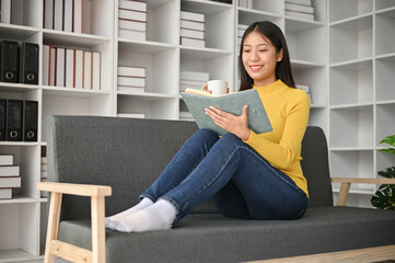 Happy young Asian woman reading book in her free time while sitting on the sofa