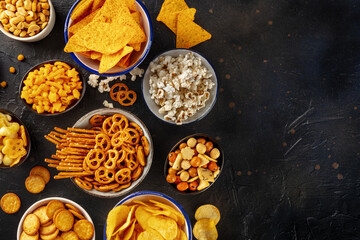 Salty snacks, party mix, overhead flat lay shot with copy space. An assortment of appetizers on a black background. Potato and tortilla chips, crackers, popcorn etc