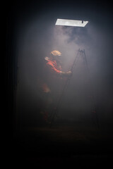 Vertical picture of firefighter is trying to climb a ladder while another firefighter is holding out his hand to help in the dark room with soft light.