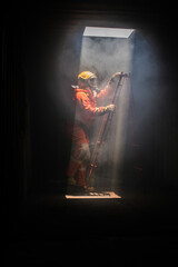 Vertical picture of firefighter is climbing a red ladder in a dark room through which light pours in from a hole above.