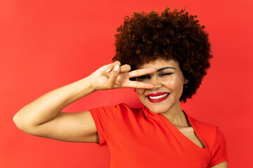 A dark-skinned smiling girl with curly afro hair posing on a red background in clothes of the same color. The young woman looks towards the camera while making the victory gesture near the eye.