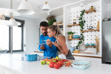 Happy young couple have fun in kitchen while preparing healthy organic food. Beautiful sports people are talking and smiling while cooking vegetarian meal in domestic kitchen at home.