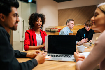 Portrait of confident African American business woman sitting at desk with laptop, looking at camera, during multicultural professional businesspeople working together on research plan in boardroom.