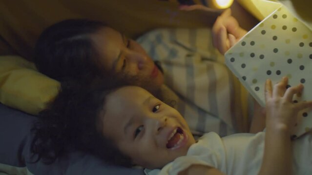 Medium close-up shot of cute Asian toddler girl smiling and playing while reading book together with mother holding flashlight under blanket on bed at night