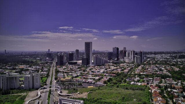 Hyperlapse video of high-rise buildings in Zapopan, Jalisco, featuring vehicle traffic on Avenida Acueducto, surrounding houses, Guadalajara city skyline, and a blue sky.