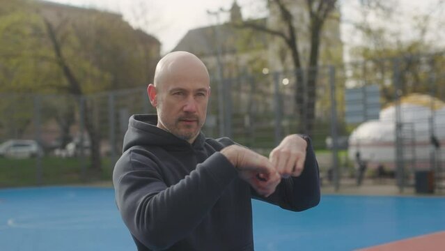 A bald man in a sportswear is exercising on a sports field. A male athlete is stretching his arms on a sports field.