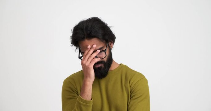 Depressed bearded young man with eyes closed shaking and touching his head in pain while standing isolated against white background
