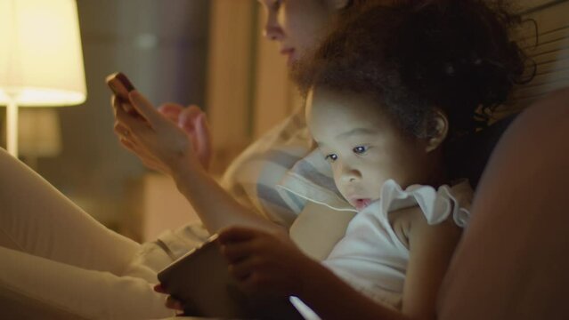 Medium close up shot of adorable Asian toddler girl watching cartoon on digital tablet while mother using phone on bed in evening at home
